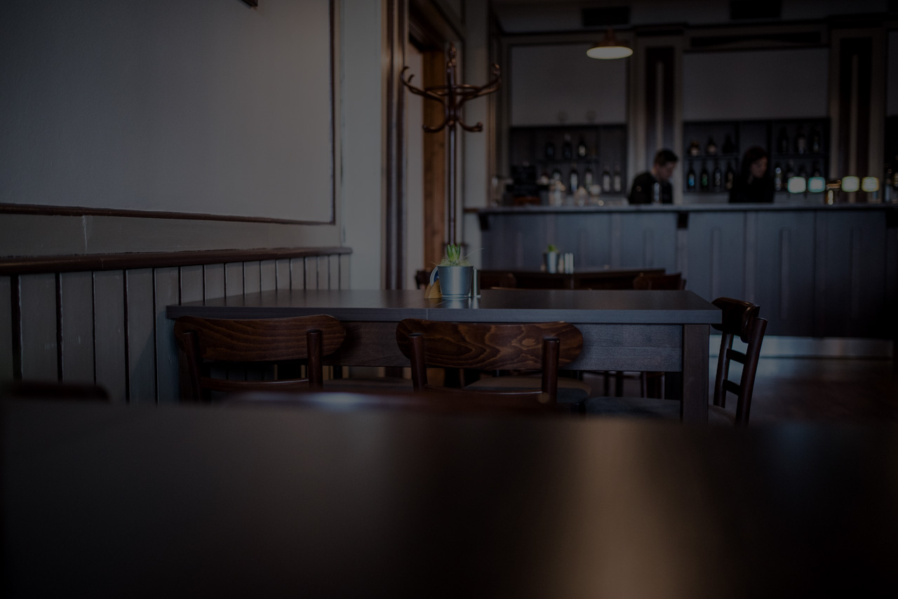 Tmavě osvětlený interiér restaurace s dřevěnými stoly a barem v pozadí